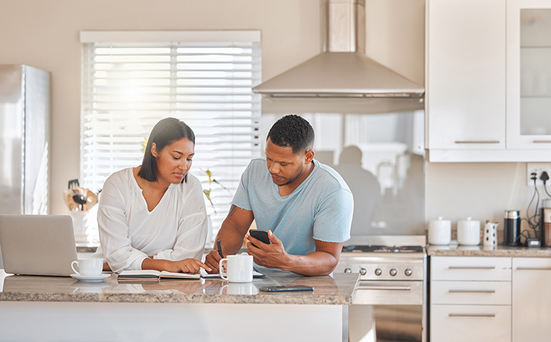 Two people ckecking their financial information while standing together in a bright kitchen with a laptop, financial journals, and their phones. 